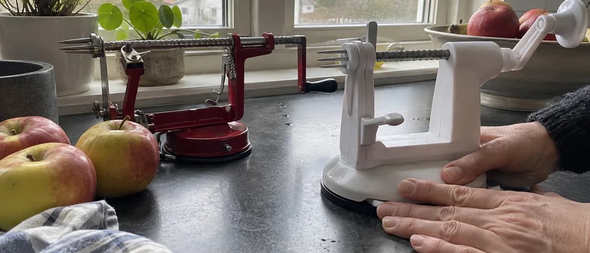 Två äppelsvarvar bredvid varandra på en köksbänk, en i metall och en i plast, medan ett par händer trycker fast sugfoten på den ena