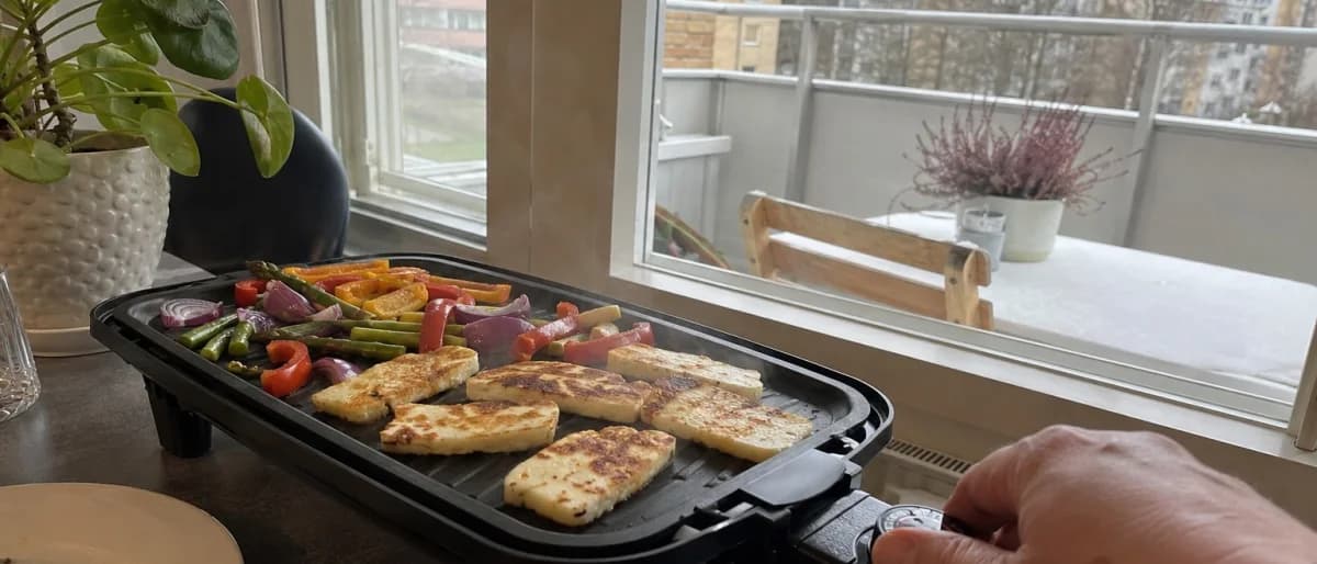 Hand justerar temperaturratten på en bordsgrill med halloumi och grönsaker, balkong synlig genom fönstret