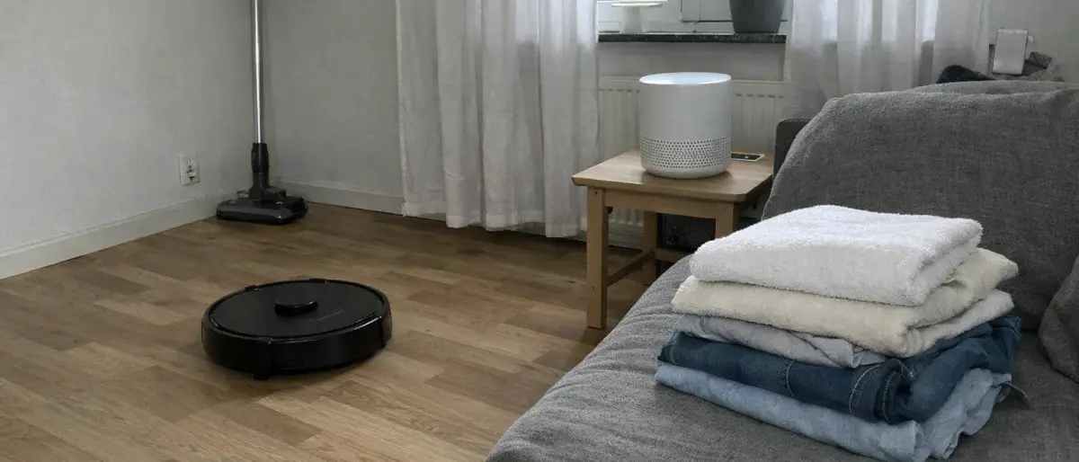 Svenskt vardagsrum med robotdammsugare på trägolv, skaftdammsugare vid väggen och vikta handdukar på soffan