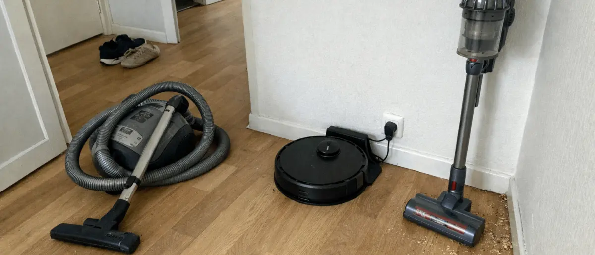 Tre dammsugare i en svensk hall — golvdammsugare, robotdammsugare vid laddstationen och skaftdammsugare i hörnet