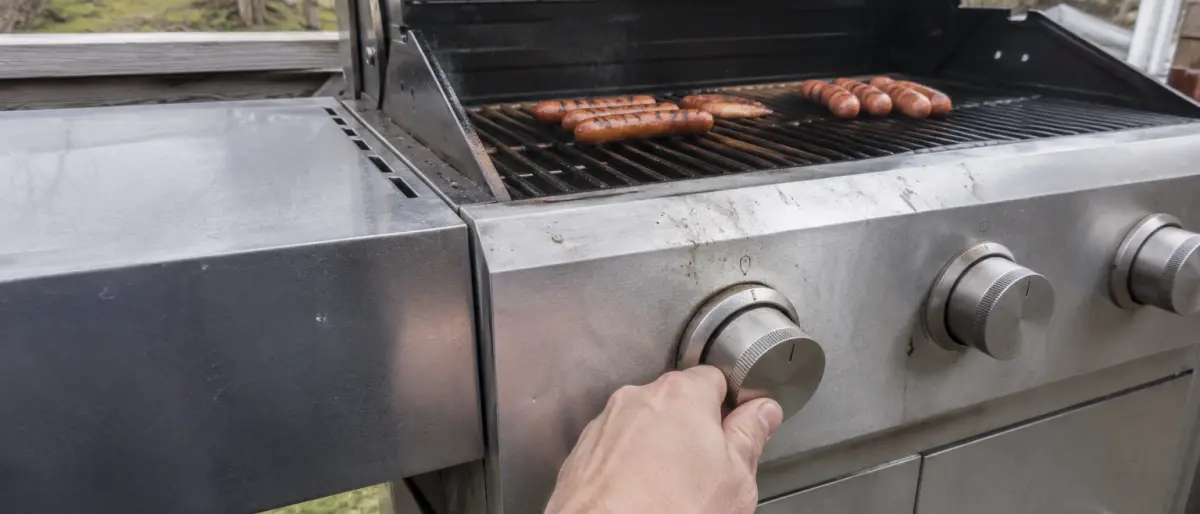 Hand som vrider på gasolgrillens vred medan korv ligger på gallret med locket öppet