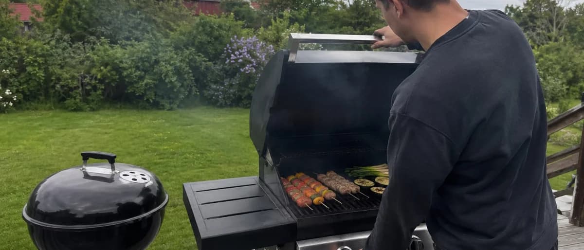 Person som lyfter på gasolgrillens lock och vänder grillspett medan en svart klotgrill står bredvid på trädäcket
