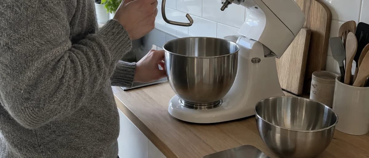 Person i grå stickad tröja håller en degkrok framför en hushållsassistent med uppfällt maskinhuvud på en köksbänk
