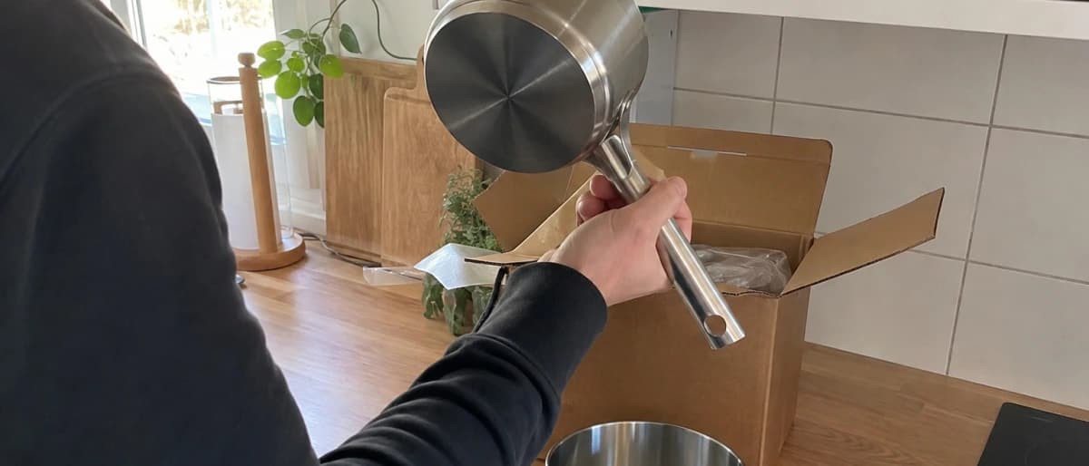 Person vid köksbänken som håller upp en kastrull och tittar på bottenplattan, med ett kastrullset delvis uppackat i en kartong bredvid