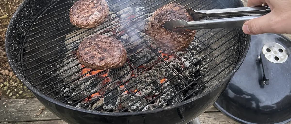 Hand med grilltång som vänder hamburgare på en kolgrill med glödande kol