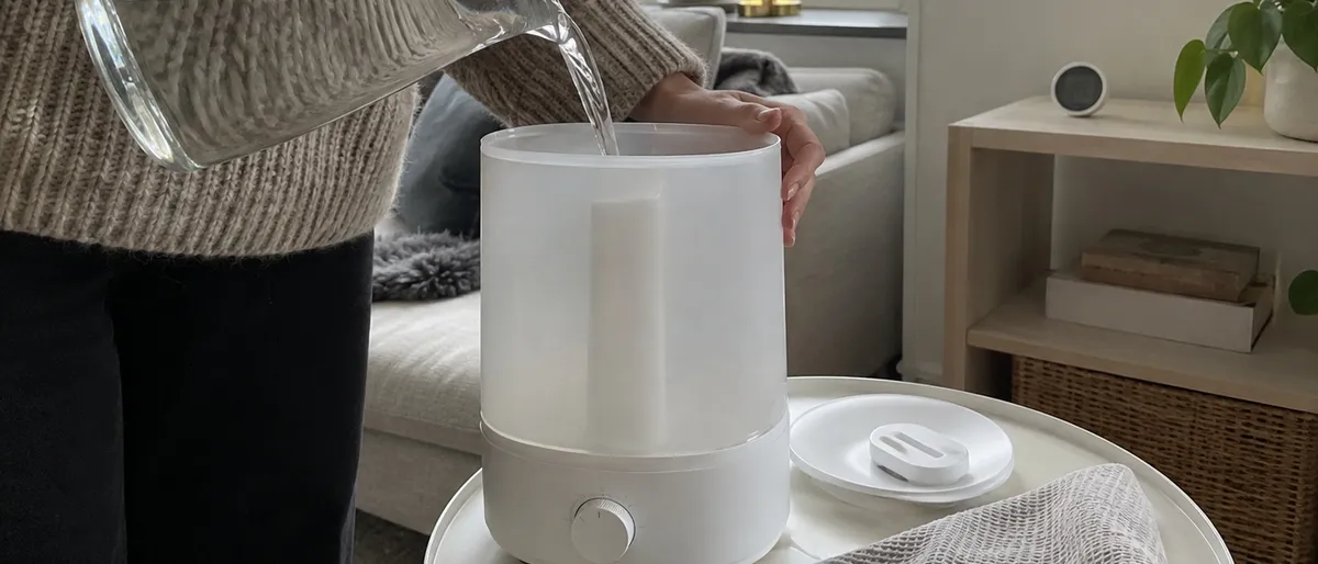 En person i stickad tröja häller vatten från en glaskanna i tanken på en vit luftfuktare som står på ett runt sidobord i ett ljust vardagsrum.