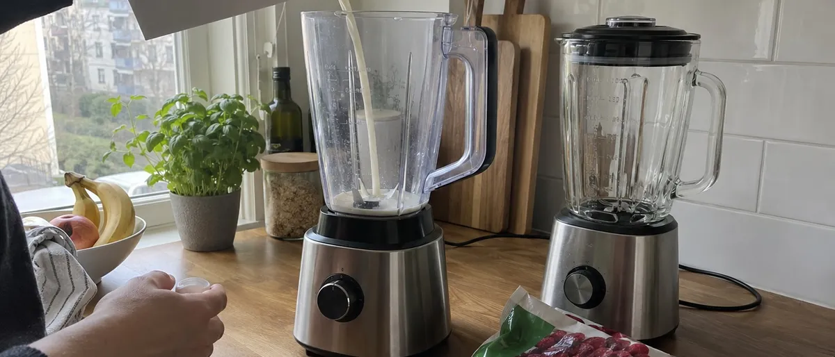 Havredryck hälls i en bordsmixer med plastkanna bredvid en mixer med glaskanna och en påse frysta hallon