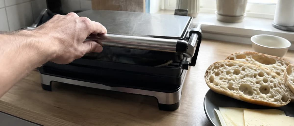 Hand som trycker ner locket på en paninigrill med bröd och ostskivor framme