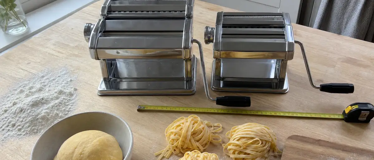 Två manuella pastamaskiner i rostfritt stål bredvid varandra på en träbänk med mjöl, pastadeg och färska tagliatelle, redo att jämföras.