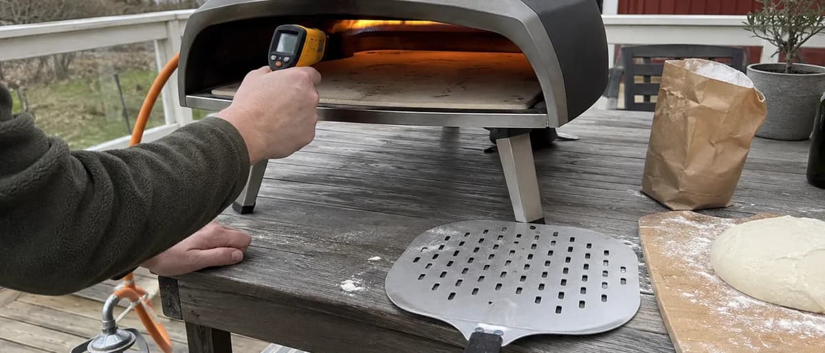 Någon mäter pizzastenens temperatur med en infraröd termometer i öppningen på en gasoldriven pizzaugn, med en hålad pizzaspade, mjöl och en degboll på trädgårdsbordet