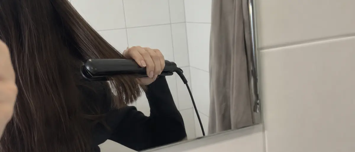 Spegelbild av en hand som plattar en hårslinga med en svart plattång i badrummet