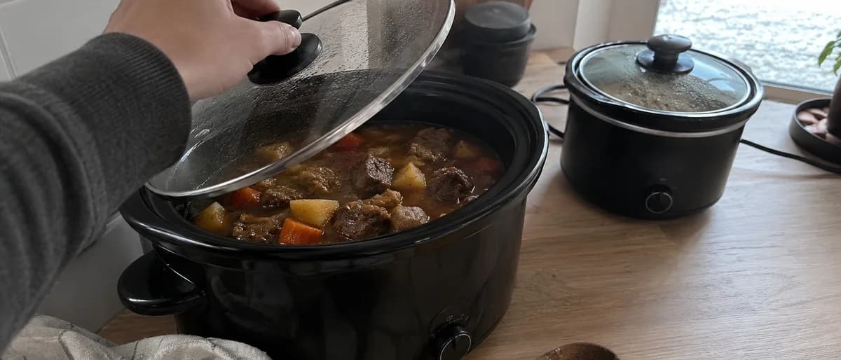 Person lyfter på glaslocket till en slow cooker med en mustig köttgryta, med en mindre slow cooker bredvid på bänken