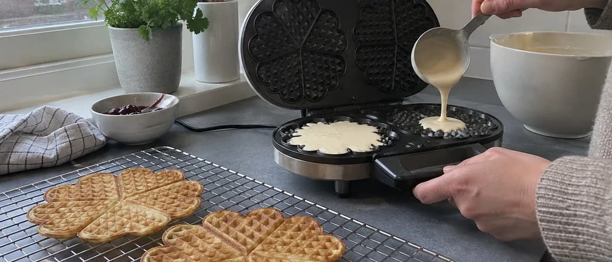 En person häller smet från en slev ner i ett öppet dubbelt hjärtvåffeljärn, med färdiga hjärtvåfflor på ett galler bredvid